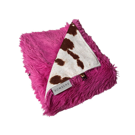 A folded bright pink faux fur blanket with a triangular white fleece insert featuring brown cow-print spots, shown on a plain white background. A small label reads BOWSERS.