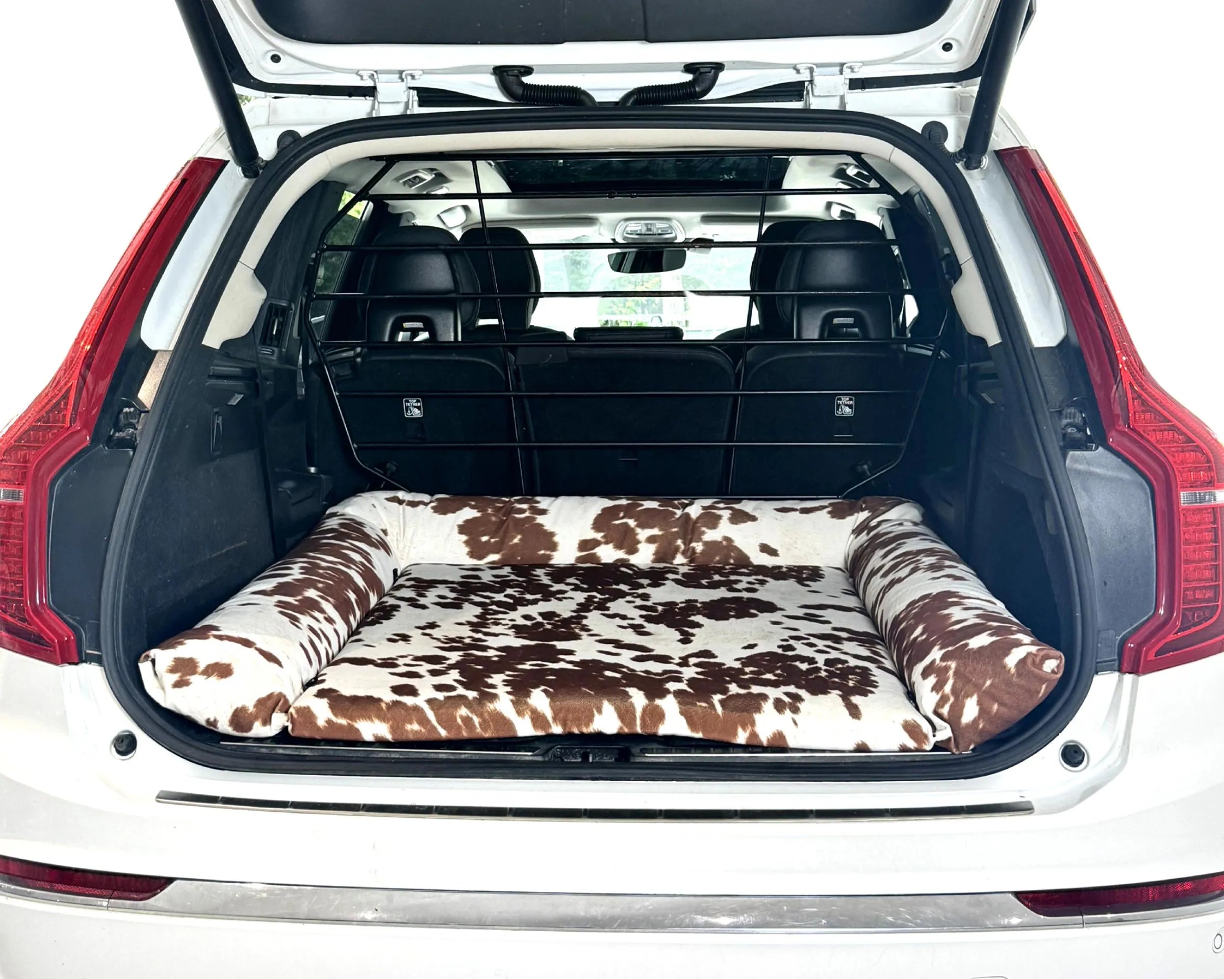 Bowsers SUV Cargo Lounger Dog Bed A car trunk pet bed with a brown and white cow print pattern, shown installed inside the open trunk of a white vehicle.