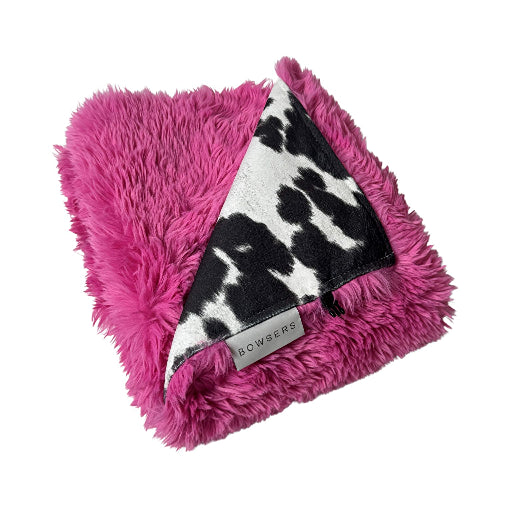 A folded plush throw blanket featuring a vibrant pink faux fur exterior and a black and white cow print interior, with a small silver 'BOWSERS' brand tag visible.