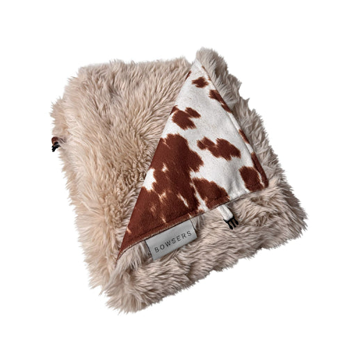 A folded faux fur blanket in a light beige/tan color, featuring a triangular patch of brown and white cow print fabric on the reverse side. A fabric label reading 'BOWSERS' is visible near the edge.