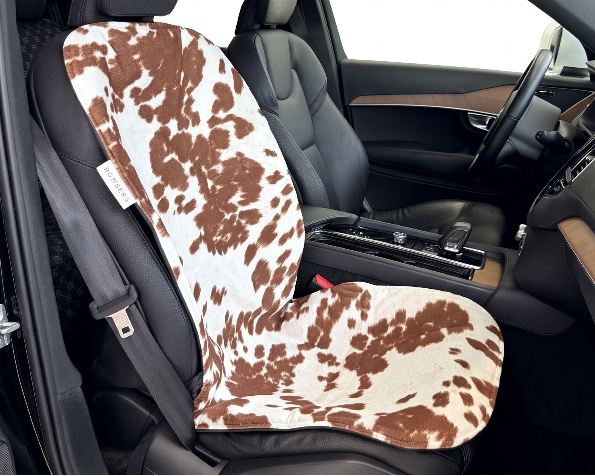 Bowsers Single Bucket Seat Cover Durango.  A car seat cover with a brown and white cow print pattern installed on a black leather car seat, shown inside a vehicle cabin.