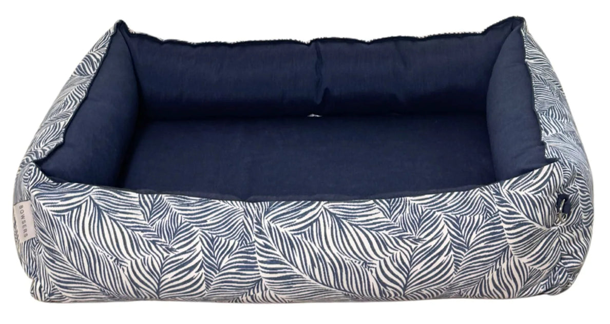A rectangular pet bed with a navy blue cushioned interior and a white side panel featuring a navy blue palm leaf pattern.