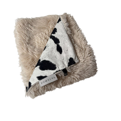 Dog Travel Blanket A square faux fur throw blanket with a beige sherpa backing and a black and white cow print reverse side, featuring a visible 'BOMBERS' brand tag.