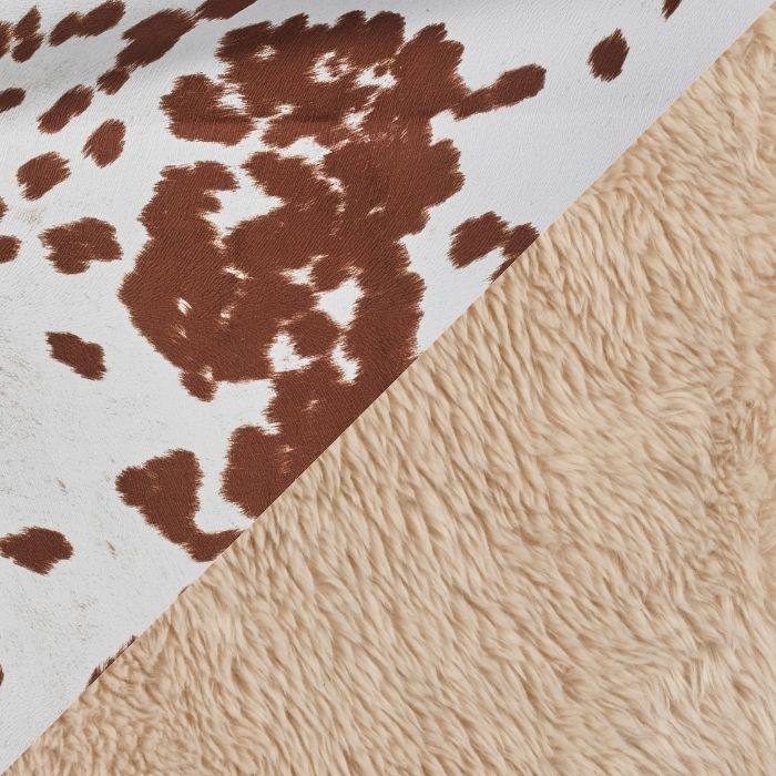 Close-up of a two-sided textile product showing a brown and white cow-print side and a soft beige sherpa fleece side, divided diagonally.